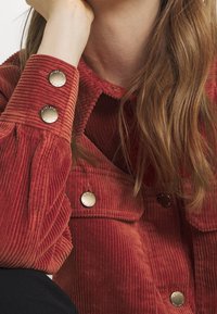 Chaqueta de pana roja con grandes botones plateados, un cuello y una tela con textura. La imagen muestra la manga y parte del cuerpo.