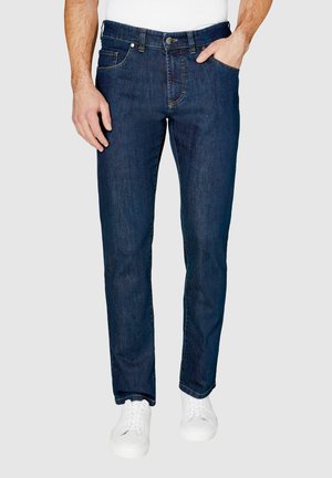 Mann trägt dunkelblaue Slim-Fit-Jeans und weiße Sneakers, eine Hand in der Tasche, vor einfachem Hintergrund.