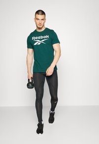 T-shirt teal da Reebok com logótipo branco, leggings pretas e sapatilhas de desporto pretas. O modelo segura um kettlebell. Fundo simples.