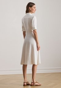 Bela lanena obleka s kratkimi rokavi, ovratnim izrezom in razširjeno krilo. Izstopajoča tekstura in preprost dizajn, skupaj z rjavimi sandali.