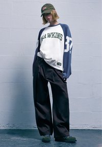 Hanorac supradimensionat alb și bleumarin cu "HAWKINS" în verde, asortat cu pantaloni largi maro și încălțăminte verde. Stilizat cu o șapcă măslinie.