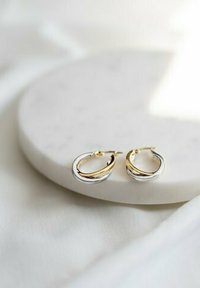 Guld- og sølvøreringe med en snoet design, der reflekterer lys. Sat på en glat hvid cirkulær overflade.