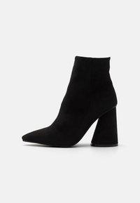 Bottine noire en daim doux, avec un bout pointu et un talon chunky de forme géométrique. Texture lisse sans ornements visibles.