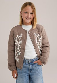 Un copil zâmbitor cu păr blond până la umeri, purtând o jachetă bej matlasată cu broderie albă, un cămașă albă și blugi.
