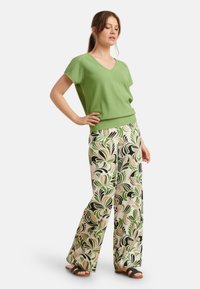 Groene, gestructureerde V-hals top gecombineerd met een broek met brede pijpen in een gedurfd, bloemig patroon in groen, zwart en beige. Er worden platte sandalen gedragen.