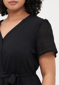 Black dress featuring short lace sleeves, a V-neckline, and a fabric belt at the waist. Smooth texture with intricate lace detailing.