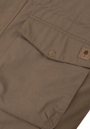 Bruine stof met een gladde textuur, gekenmerkt door een klepzak die is bevestigd met een ronde knop en een klein boomlogo op een tankleurig patch.