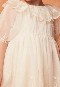 Robe blanc cassé avec manches bouffantes transparentes, encolure à volants et broderies florales. Jupe évasée avec taille froncée et texture lisse.