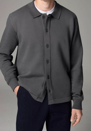 Hombre con cárdigan gris oscuro abotonado sobre camiseta blanca de cuello redondo y pantalones oscuros, posando frente a un fondo gris liso.