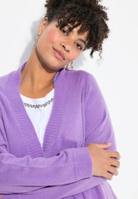 Cardigan viola con scollo a v, polsini e orlo a coste. Tessuto morbido, indossato sopra una maglietta bianca con una collana d'argento.