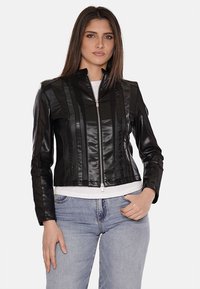 Giacca in finta pelle nera con design a righe verticali, chiusura con zip, taglio corto e colletto rialzato, abbinata a una maglietta bianca e jeans blu.