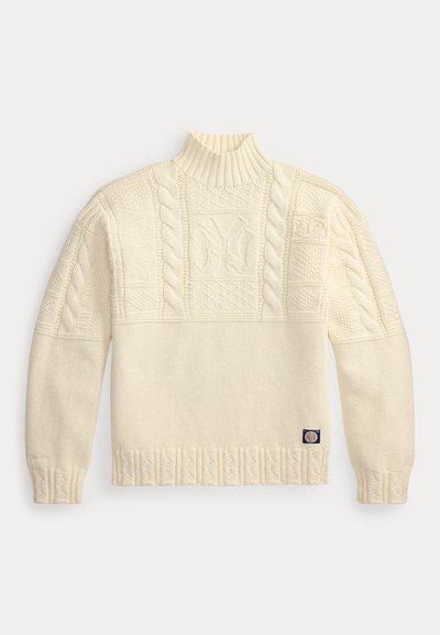 Πλεκτό πουλόβερ σε κρεμ χρώμα με υψηλό ριμπ λαιμό, σχέδια με πλέξη κόμπος και ένα μικρό υφασμάτινο ταμπελάκι κοντά στο τελείωμα.