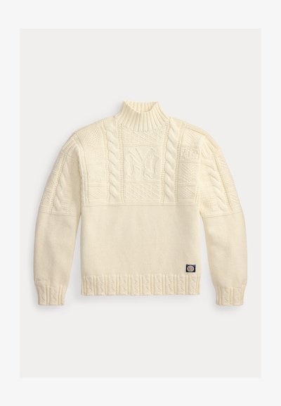 Πλεκτό πουλόβερ σε κρεμ χρώμα με υψηλό ριμπ λαιμό, σχέδια με πλέξη κόμπος και ένα μικρό υφασμάτινο ταμπελάκι κοντά στο τελείωμα.
