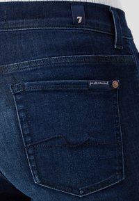 Donkerblauwe denim jeans hebben een klassiek vijf-pocket ontwerp, zichtbare stiksels en een label met de tekst "for all mankind" op het achterzakje.