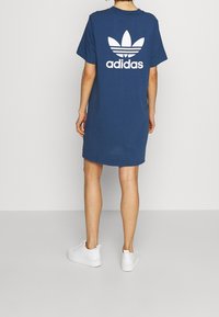 Námořnická modrá bavlněná šaty ve stylu trička s velkým bílým logem Adidas a grafikou trojlístku na zádech, doplněné o bílé tenisky.