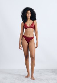 Bordowy koronkowy biustonosz typu bralette i pasujące majtki bikini, charakteryzujące się klasycznym wzornictwem z trójkątnymi kształtami i miękkim teksturowanym materiałem.