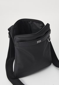 Black canvas shoulder bag with a zip closure, smooth texture, BOSS logo on the front, and an adjustable strap. Interior is lined.