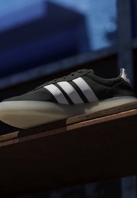 Scarpa da ginnastica Adidas bianca e nera con tre strisce bianche che riposa su una superficie in legno contro uno sfondo blu scuro sfocato.