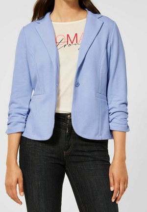 Kobieta ubrana w jasnoniebieską dopasowaną marynarkę na białej koszulce z nadrukiem oraz czarne, wysokie jeansy z widocznymi guzikami.