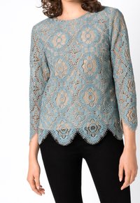 Spetstopp med blommönster i ljusblått, med trekvartslång ärm och scallopad nederkant, parat med svarta byxor.