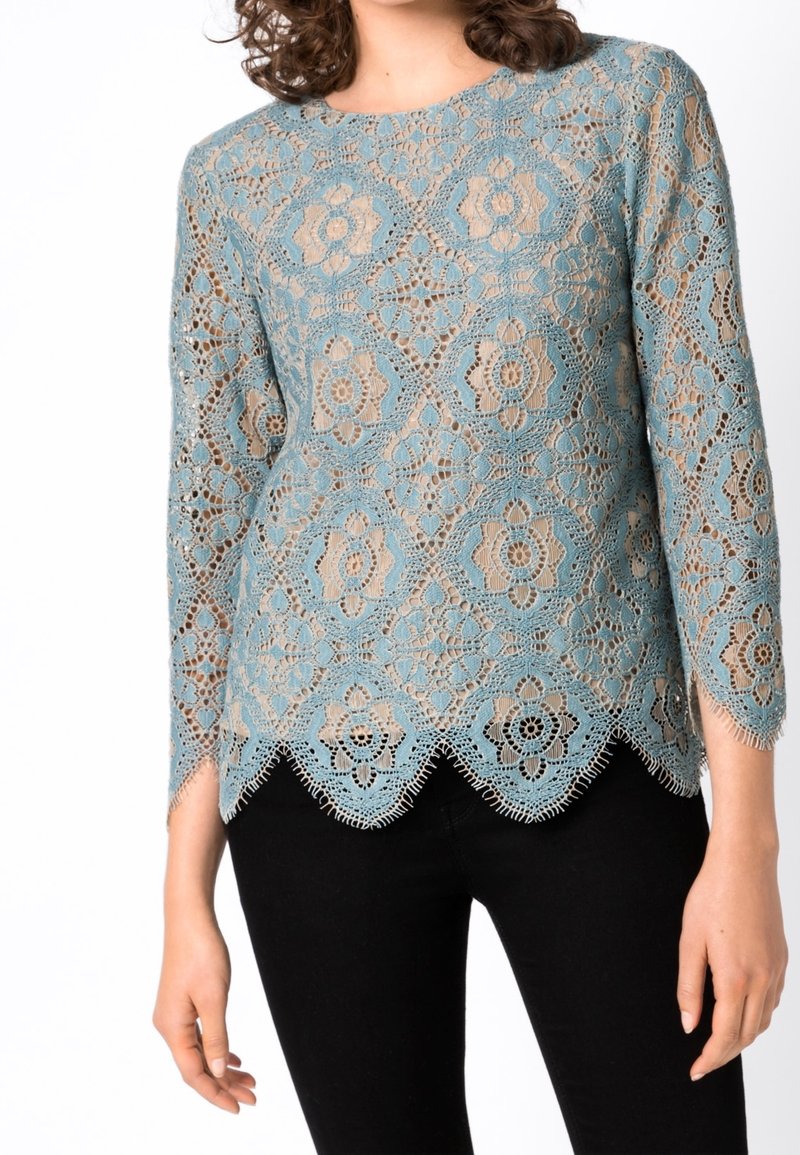 Spetstopp med blommönster i ljusblått, med trekvartslång ärm och scallopad nederkant, parat med svarta byxor.
