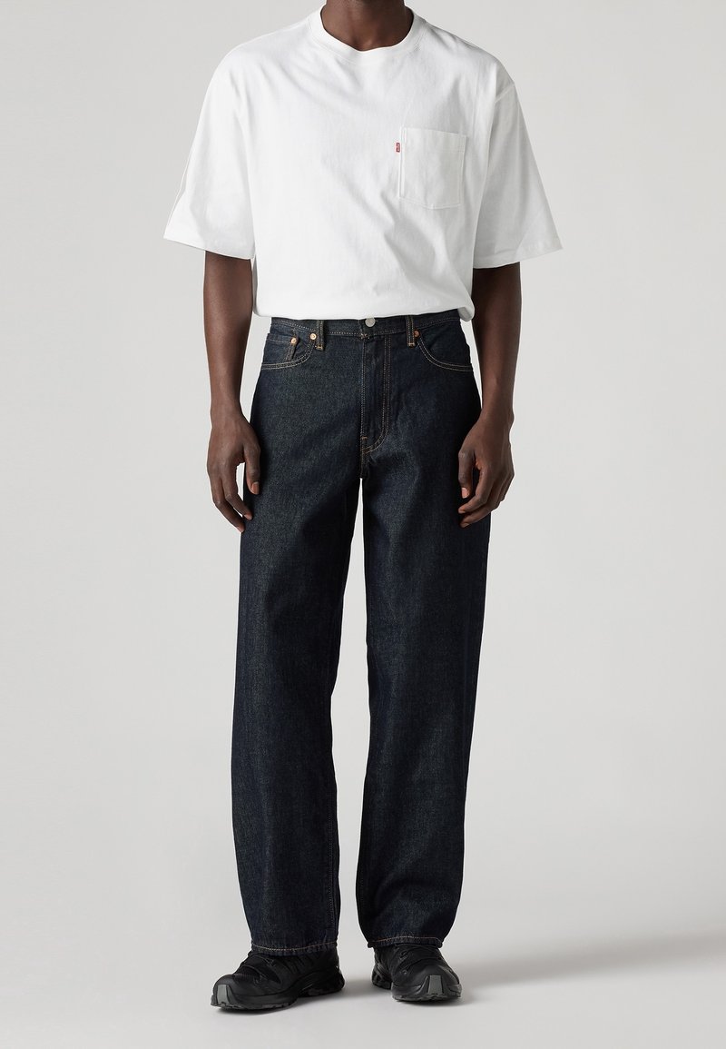 Personne portant un T-shirt blanc à manches courtes avec une poche poitrine, un jean bleu foncé ample et des chaussures noires, debout sur un fond uni clair.