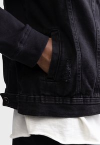 Mano en el bolsillo de una chaqueta de mezclilla negra con detalles deshilachados, llevada sobre una camisa blanca y la manga de un sudadera oscura.