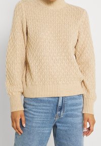 Beige Strickpullover mit gerippten Bündchen und Saum, getragen mit hellblauen, hochtaillierten Jeans von einer stehenden Person.