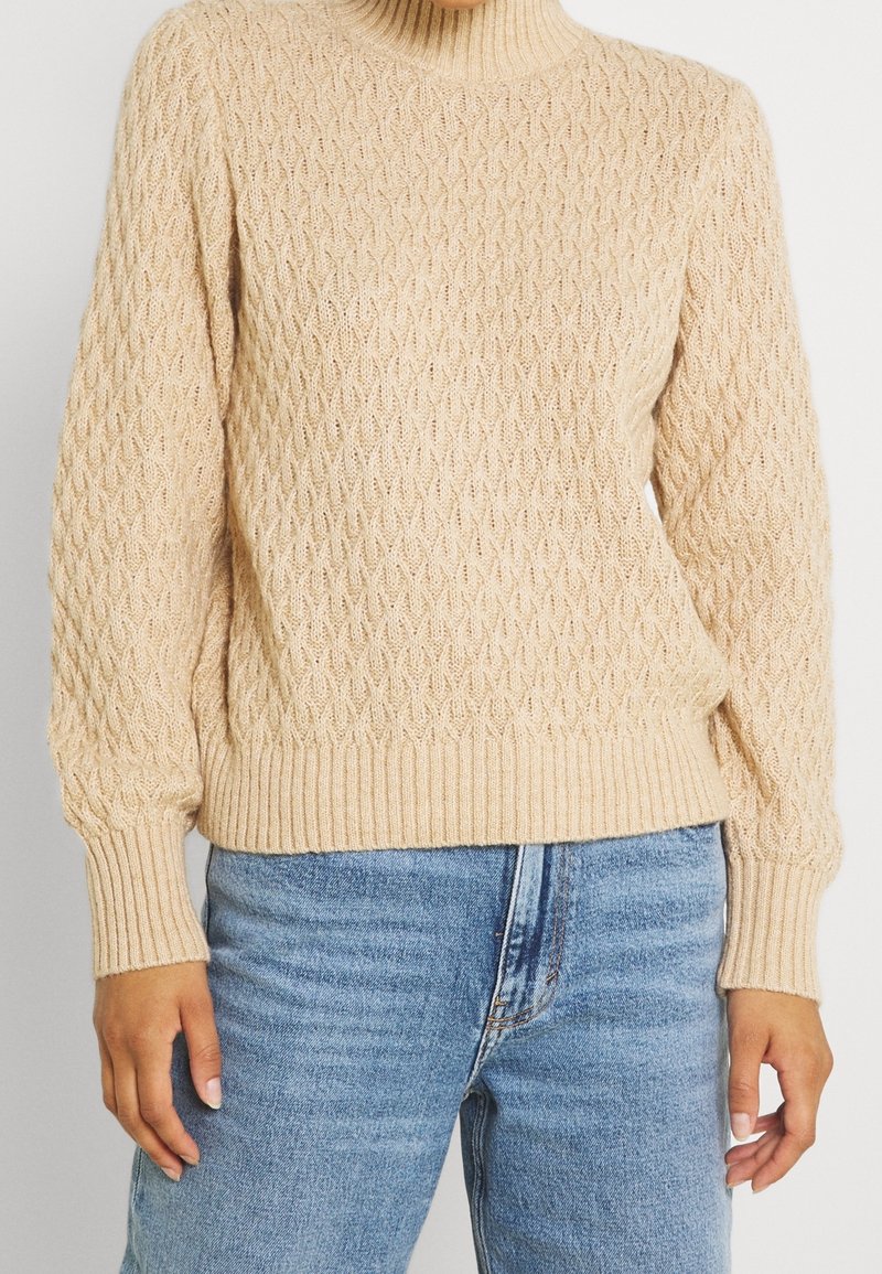 Beige Strickpullover mit gerippten Bündchen und Saum, getragen mit hellblauen, hochtaillierten Jeans von einer stehenden Person.