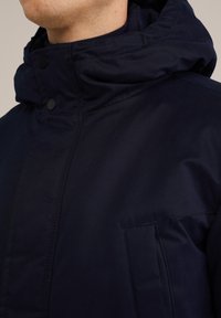 Tamsiai mėlynas pūkinis švarkas su aukštu apykakle, kapišonu ir spaudimo mygtukų užsegimu; turi priekinę kišenę ir lygią, matinę apdailą.