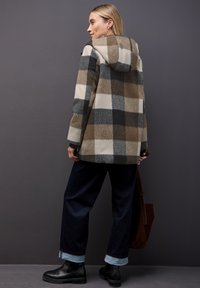 Kabát z vlněné směsi s károvaným vzorem v hnědé, béžové a šedé barvě. Má kapuci a dlouhé rukávy. Kombinován s tmavými džínami a černými kotníkovými botami.