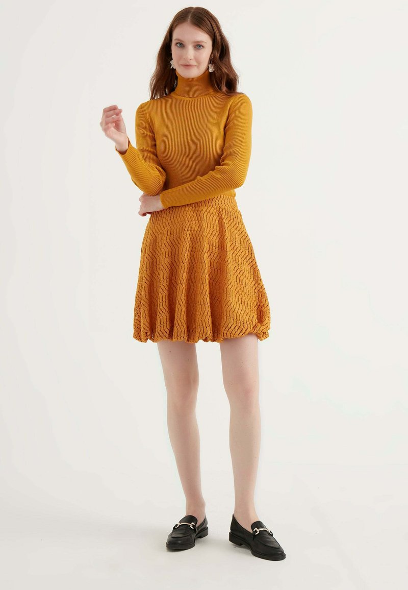 Maglione giallo senape a coste con collo alto abbinato a una gonna A-line texturizzata con motivo a zigzag. Modello con mocassini neri.