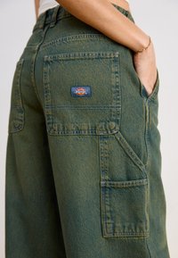 Pantaloni cargo din denim verde, având un buzunar posterior mare, cusături întărite și o etichetă de logo vizibilă pe partea dreaptă.