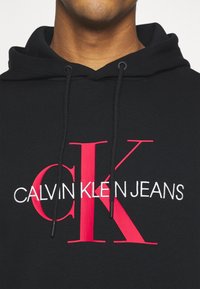 Felpa nera con cappuccio caratterizzata da un grande logo rosso "CK" e testo bianco "CALVIN KLEIN JEANS". Realizzata in tessuto morbido con dettaglio alla coulisse.
