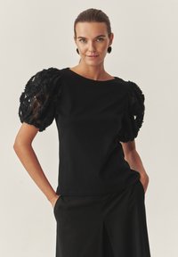 Fekete felső virágos csipkés puffos ujjakkal, kerek nyakkivágással, testhezálló fazonnal, fekete bő szárú nadrággal kombinálva. Egységesen sima textúrájú.