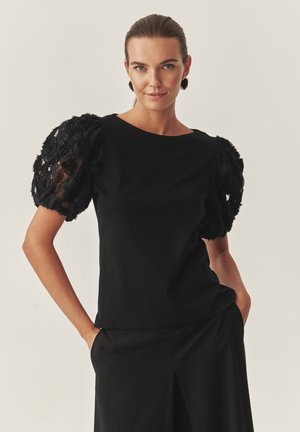 Must särk lillelise pitsiga lõuendvarrukatega, ümar kaelus, keha järgiva lõikega, kombineeritud mustade laiade pükstega. Üldine pind on sile.