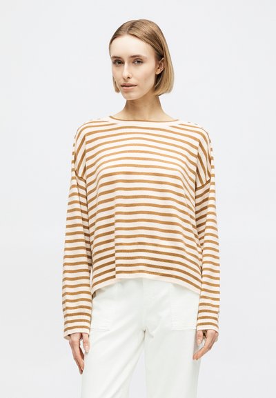Femme aux cheveux blonds courts portant un haut à manches longues beige et blanc à rayures et un pantalon blanc, debout devant un fond uni.