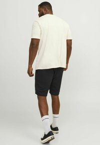 T-shirt beige chiaro a maniche corte, abbinata a pantaloni corti scuri. Presenta una vestibilità classica e un design pulito. Sneakers nere a contrasto e calzini bianchi.