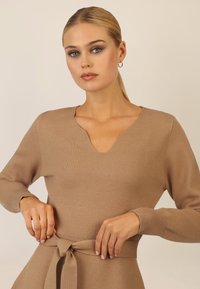 Beige gestrickter Pullover mit V-Ausschnitt, langen Ärmeln und einem Knotenbund in der Taille. Der Stoff hat eine glatte Textur und eine figurbetonte Silhouette.