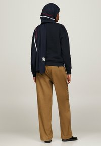 Sudadera marina, pantalones anchos beige y una bufanda a juego con rayas rojas y blancas, con un detalle de logo en el extremo de la bufanda.