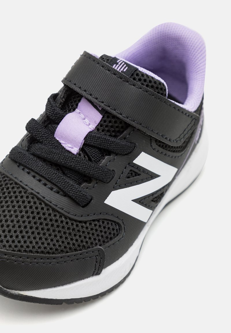 Zapatilla para niños pequeña en negro y morado con suela blanca, parte superior de malla, tira de velcro, cordones negros y logo "N" blanco en el lateral.