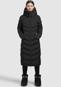 Ilgas juodas pūkinis paltas su zigzago raštu, aukštu apykakle, priekiniu užtrauktuku ir šoninėmis kišenėmis. Dėvimas su juodomis leginsomis ir šiltomis pilkomis avalynėmis.