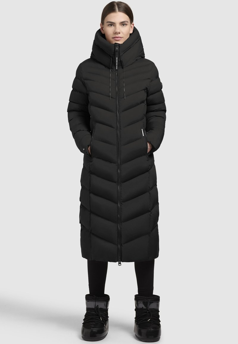 Ilgas juodas pūkinis paltas su zigzago raštu, aukštu apykakle, priekiniu užtrauktuku ir šoninėmis kišenėmis. Dėvimas su juodomis leginsomis ir šiltomis pilkomis avalynėmis.