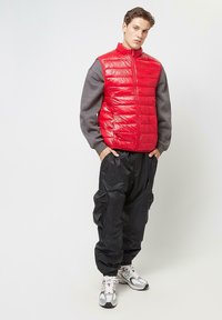 Red puffer vest with horizontal quilted pattern, gray sleeves, and zip closure. Paired with black cargo pants and white athletic shoes.
