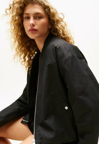 Czarna kurtka bomberka wykonana z gładkiego, wodoodpornego materiału. Posiada zamek z przodu, prążkowany kołnierz oraz kieszenie boczne. Prosty i funkcjonalny design.