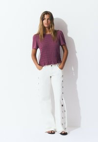 Haut smocké violet à manches courtes associé à un pantalon blanc à jambes larges avec des boutons sur les côtés et des sandales noires à glisser. Fond lumineux.