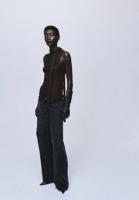 Top brown fără mâneci, cu un design ajustat și detaliu cu nod, asortat cu blugi de denim închis, cu picioare largi și mănuși din piele neagră. Fundal neutru.