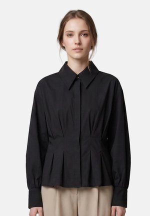 Jeune femme aux cheveux châtain clair portant un chemisier noir à manches longues boutonné et un pantalon beige, debout devant un fond uni.
