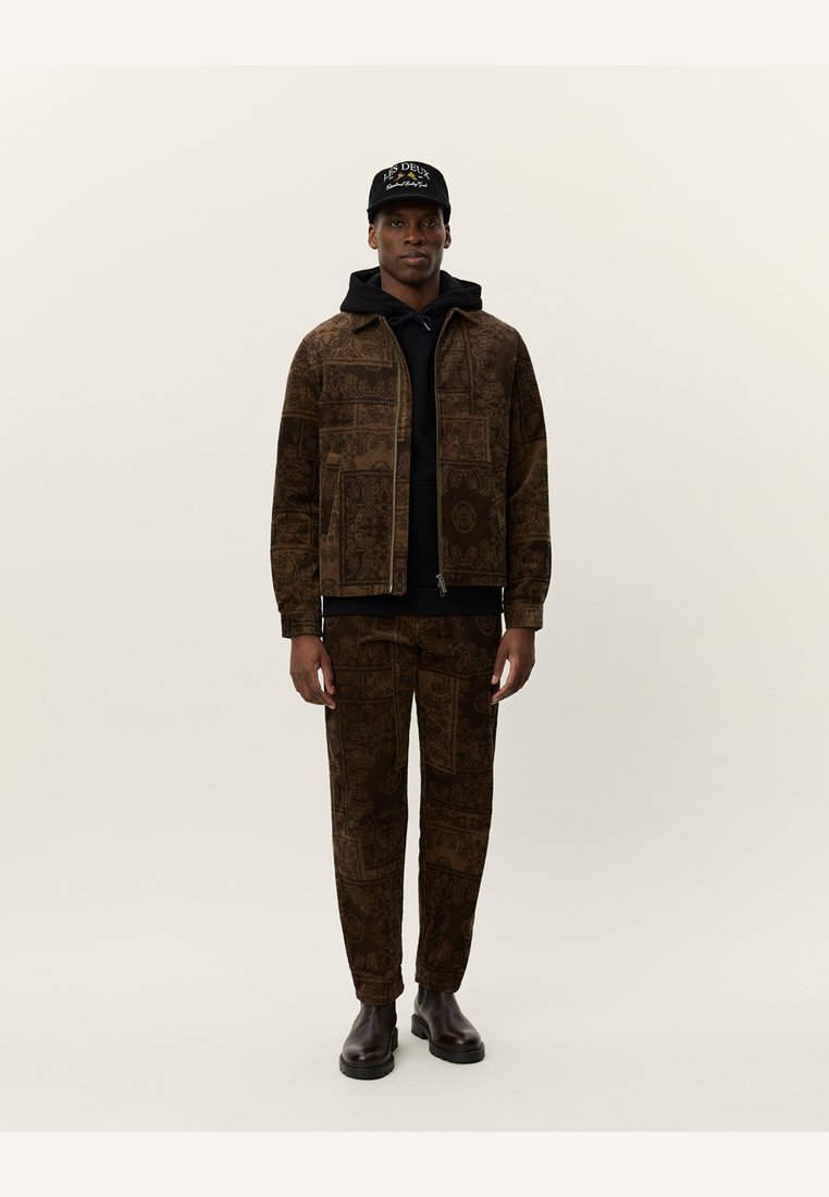 Brown patterned jacket and pants set, featuring detailed designs. Worn over a black hoodie, complemented by a black cap and brown boots.