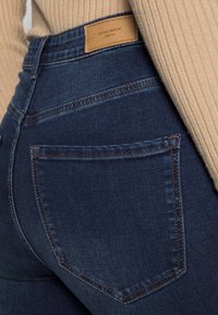 Tummansiniset farkut, joissa on sileä pinta, takaosassa takatasku ja kontrastivärinen ommel. Merkkilogo näkyvissä vyötäröllä.